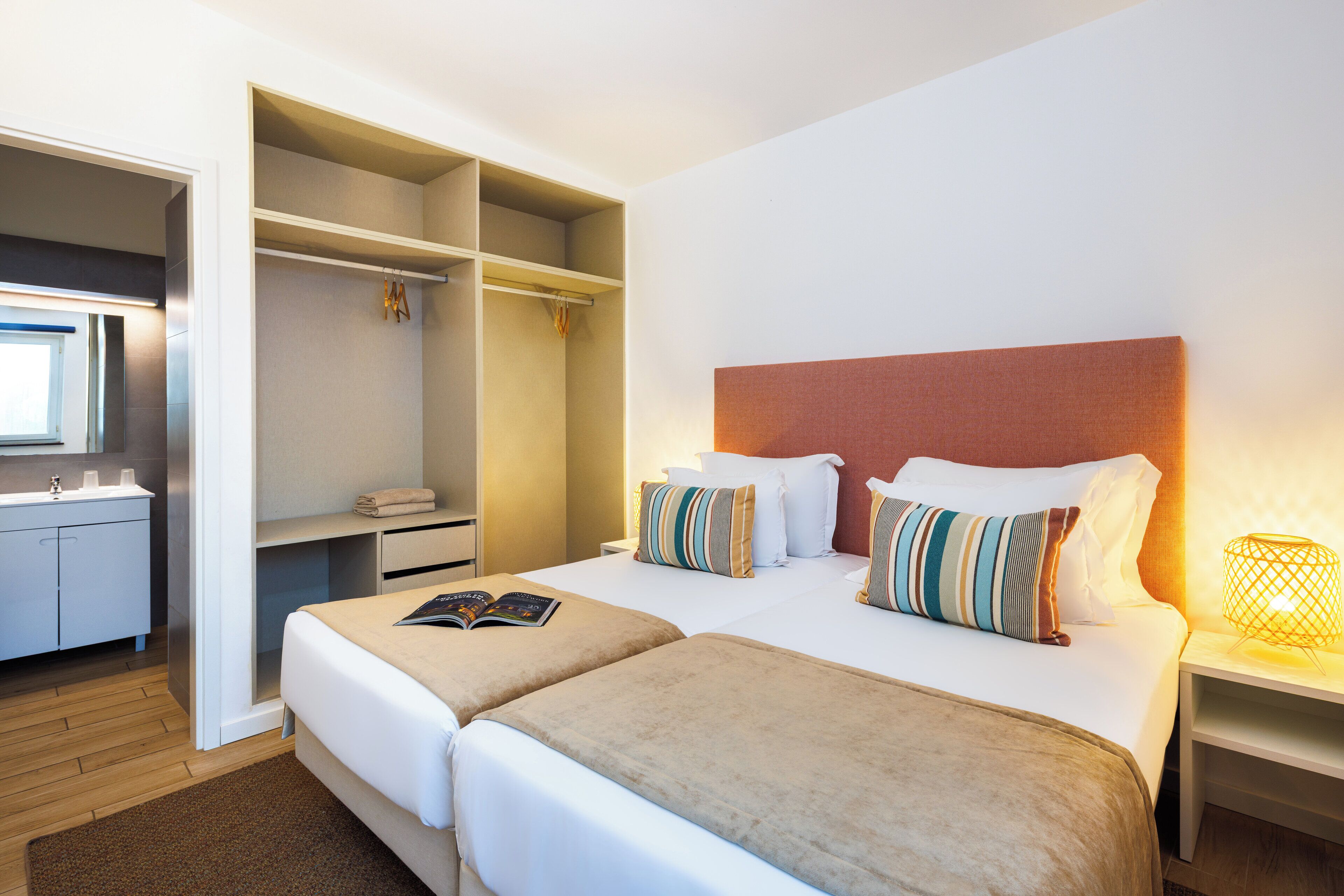 Una habitación de hotel con dos camas, un armario, un baño y una mesita de noche con lámpara.