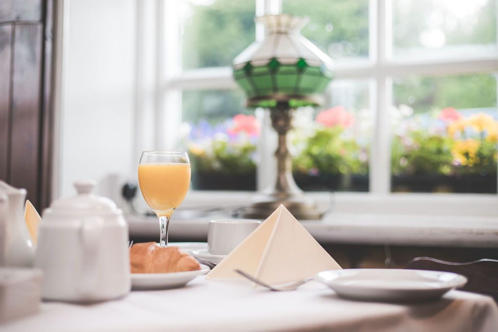 Et morgenbord med et glas appelsinjuice, en croissant og en pyramideformet serviet på et bord ved et vindue med udsigt til blomster.