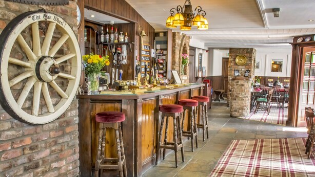 Uma área de bar com uma grande roda de madeira, banquetas e diversos frascos e copos nas prateleiras.