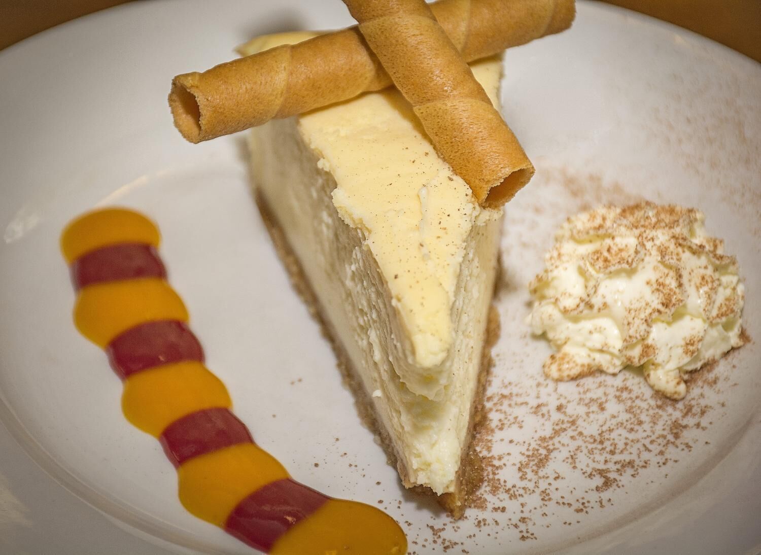 Una porción de pastel de queso con un rollo de galleta encima, una cucharada de crema batida y salsa decorativa sobre un plato.