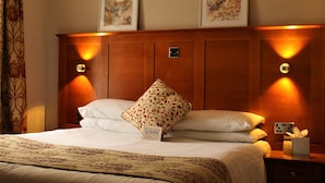 Quarto de hotel com uma cama grande, cabeceira de madeira, duas luminárias fixadas na parede, mesa de cabeceira com caixa de lenços e duas fotos emolduradas na parede.