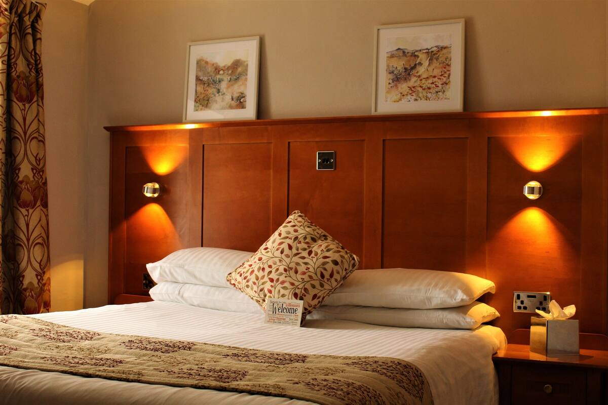 A hotel room with a large bed, a wooden headboard, two wall-mounted lamps, a bedside table with a tissue box, and two framed pictures on the wall.