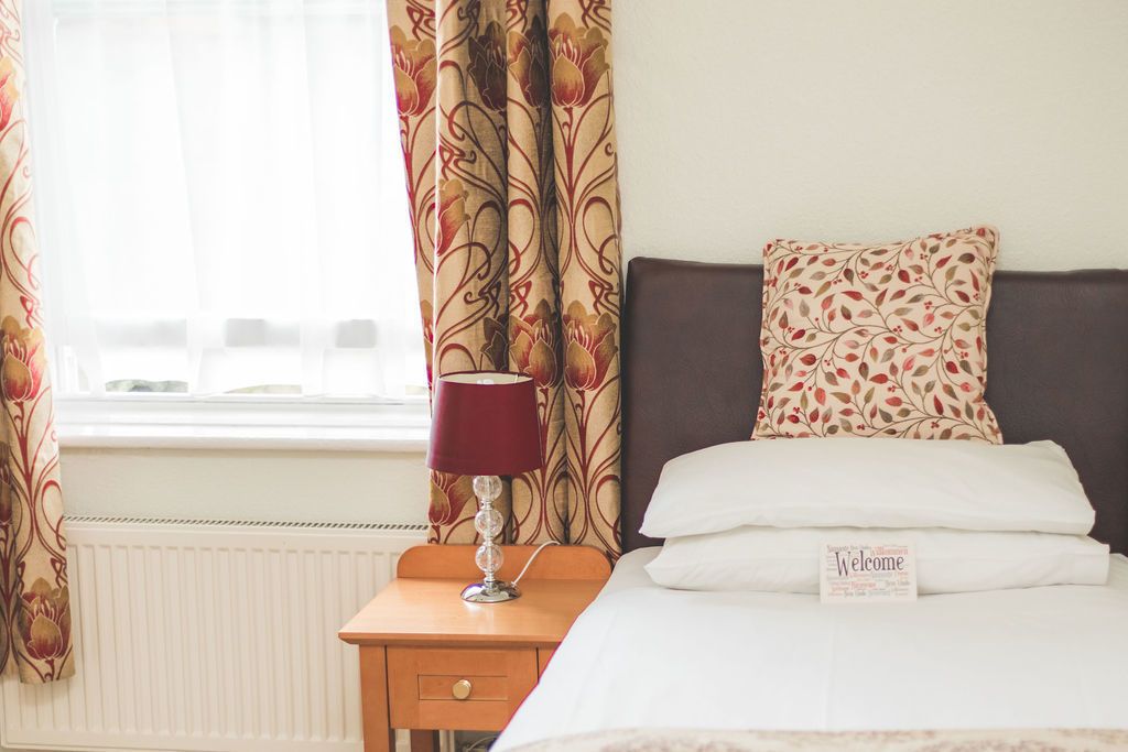 A hotel room with a bed, a nightstand with a lamp, a window with curtains, and a welcome sign on the bed.