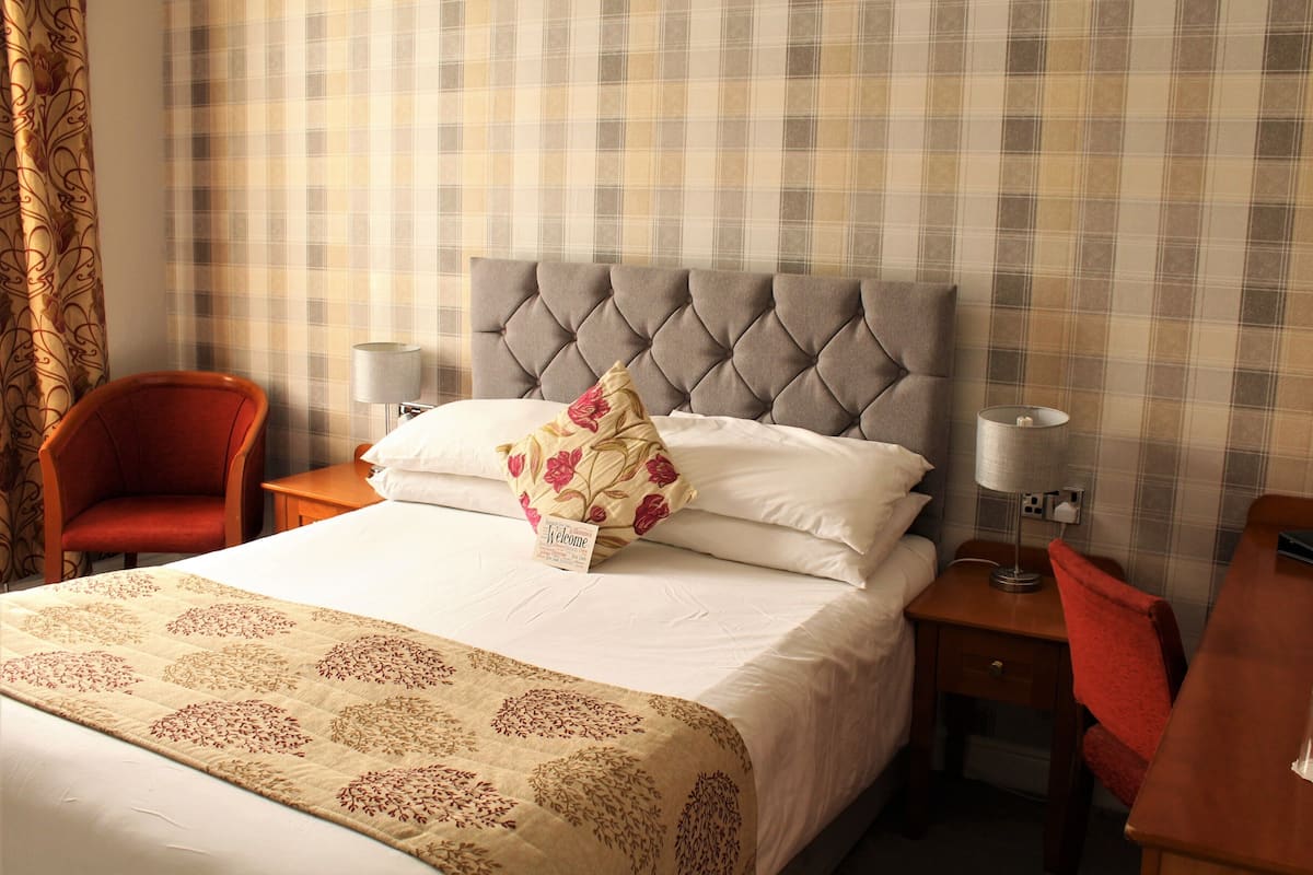A hotel room with a tufted headboard, a floral-patterned bedspread, a red armchair, bedside lamps, and a wooden nightstand.