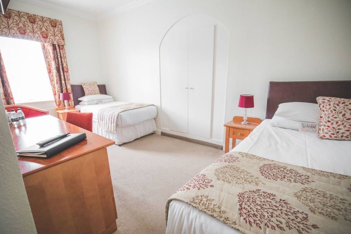 A hotel room with a desk, two beds, a wardrobe, and a window with curtains.