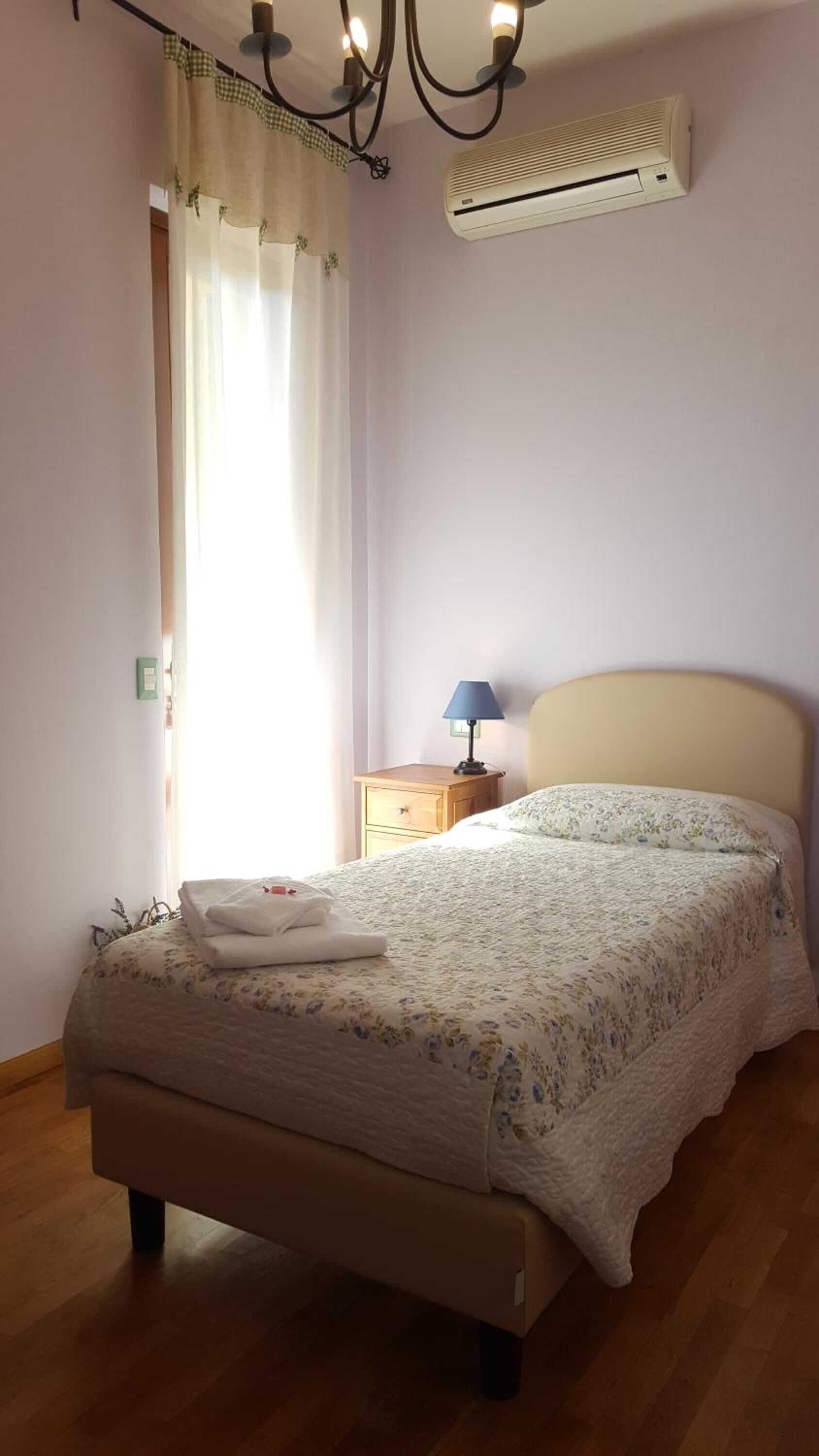 Ein Schlafzimmer mit Bett, Nachttisch, Lampe, Klimaanlage und einem Fenster mit Vorhängen.