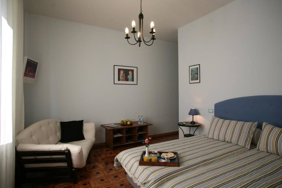 A bedroom with a bed, a chair, a nightstand, a lamp, and a chandelier.