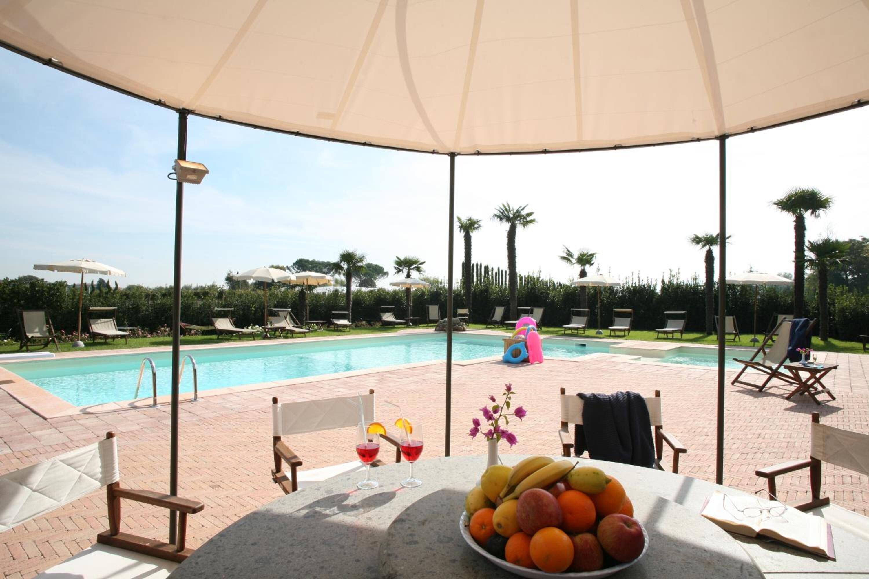 A poolside area with lounge chairs, a table with fruit and drinks, and a view of the pool and palm trees.