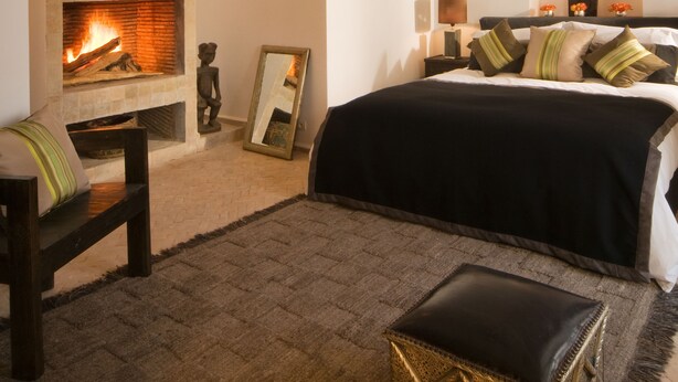 Un dormitorio con chimenea, una cama con colcha blanco y negro, un taburete pequeño y una lámpara colgante.