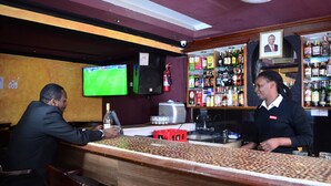 A bar with a wooden counter, a television displaying a sports game, and shelves stocked with various bottles.