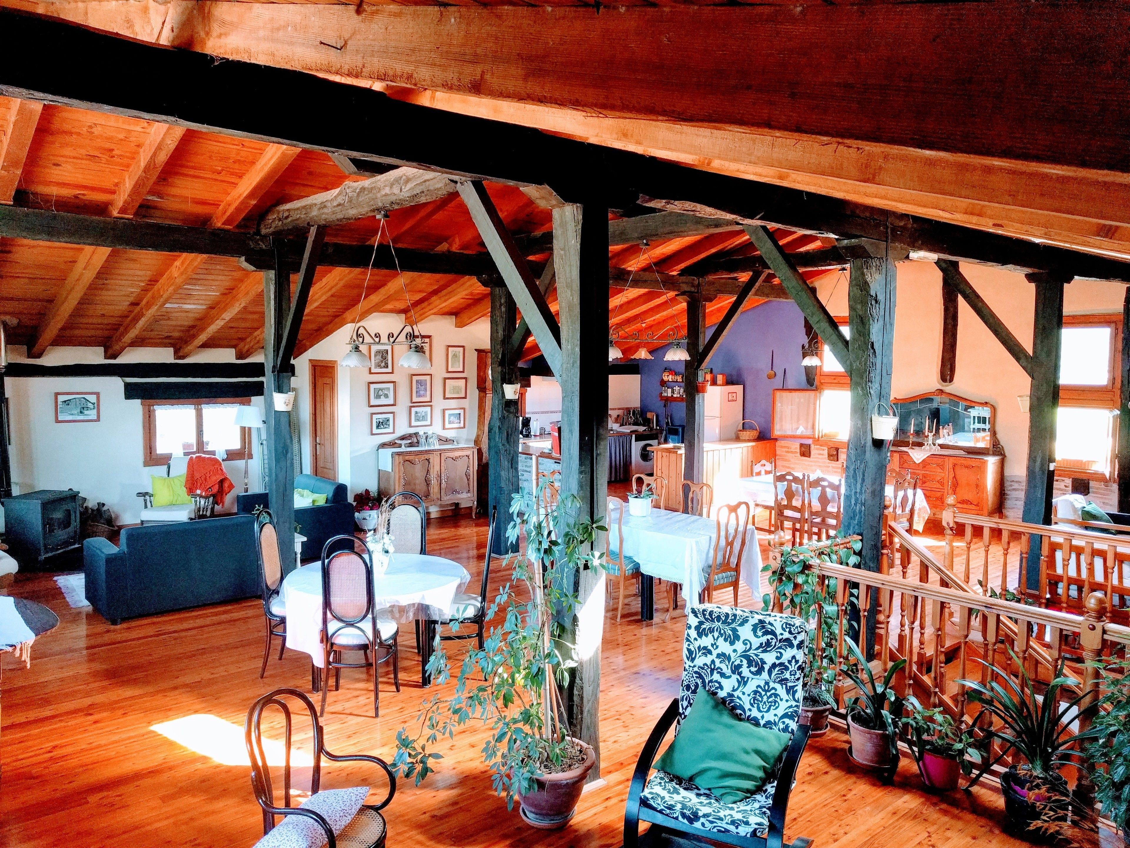 A spacious loft with high ceilings, exposed wooden beams, and a mix of seating areas including a dining table and chairs, a sofa, and a chair with a patterned cushion.