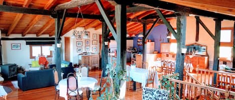 A spacious loft with high ceilings, exposed wooden beams, and a mix of seating areas including a dining table and chairs, a sofa, and a chair with a patterned cushion.