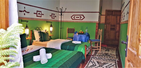 Ein Zimmer mit zwei Betten, einem Holzstuhl, einem Tisch mit Früchten und einem gemusterten Teppich.