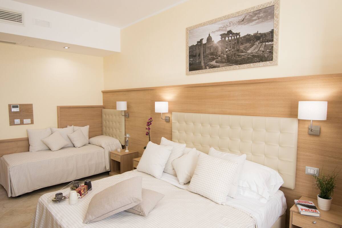Habitación de hotel con dos camas, una cama grande con cabecero capitonado y una cama más pequeña con cabecero de madera. Hay lámparas fijadas a la pared, un cuadro enmarcado y una mesita con una planta y un libro.