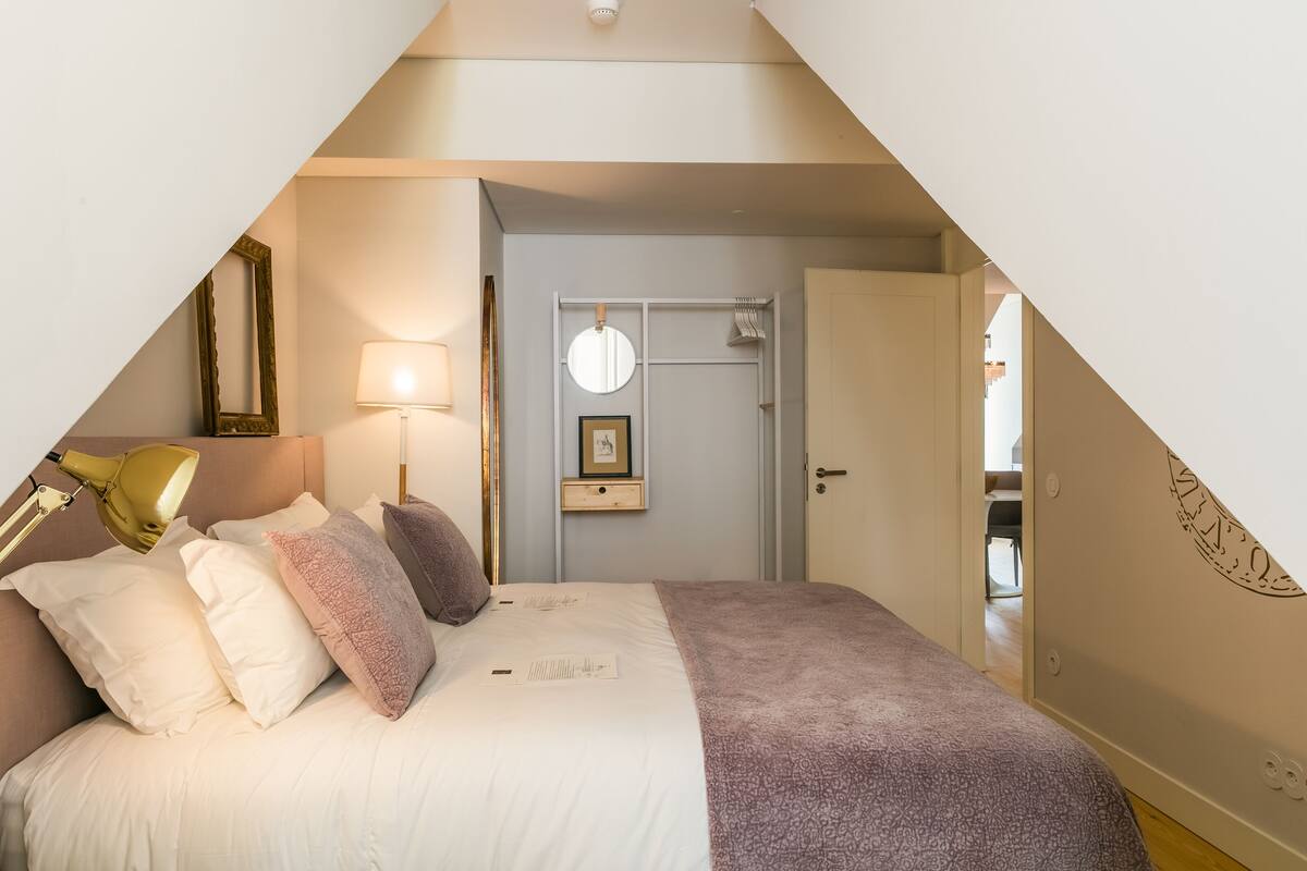 A bedroom with a sloped ceiling, a bed with white and pink bedding, a bedside lamp, a framed picture, and a door leading to another room.