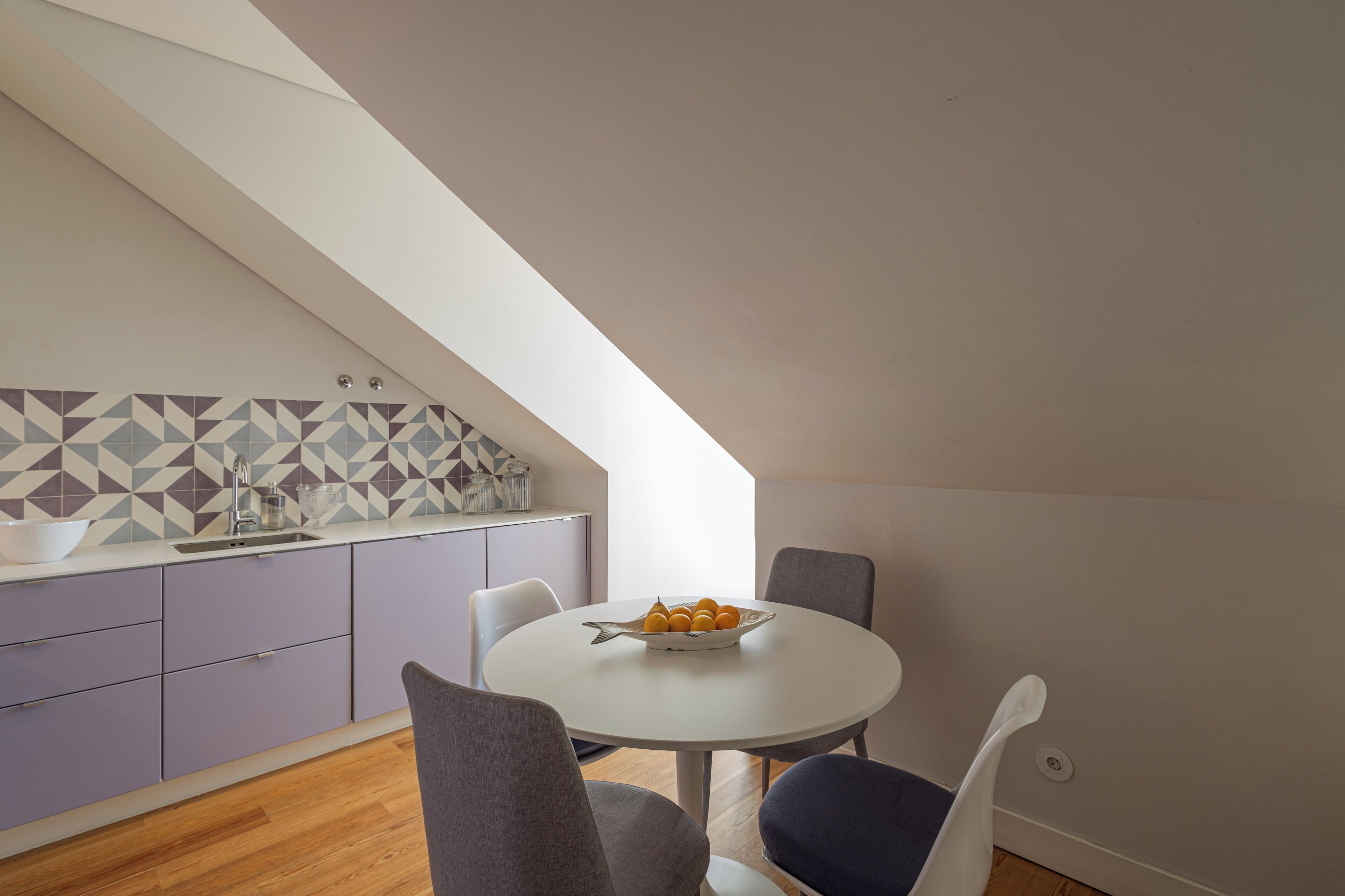 A modern kitchen with a geometric backsplash, a white dining table with chairs, and a bowl of fruit on the table.