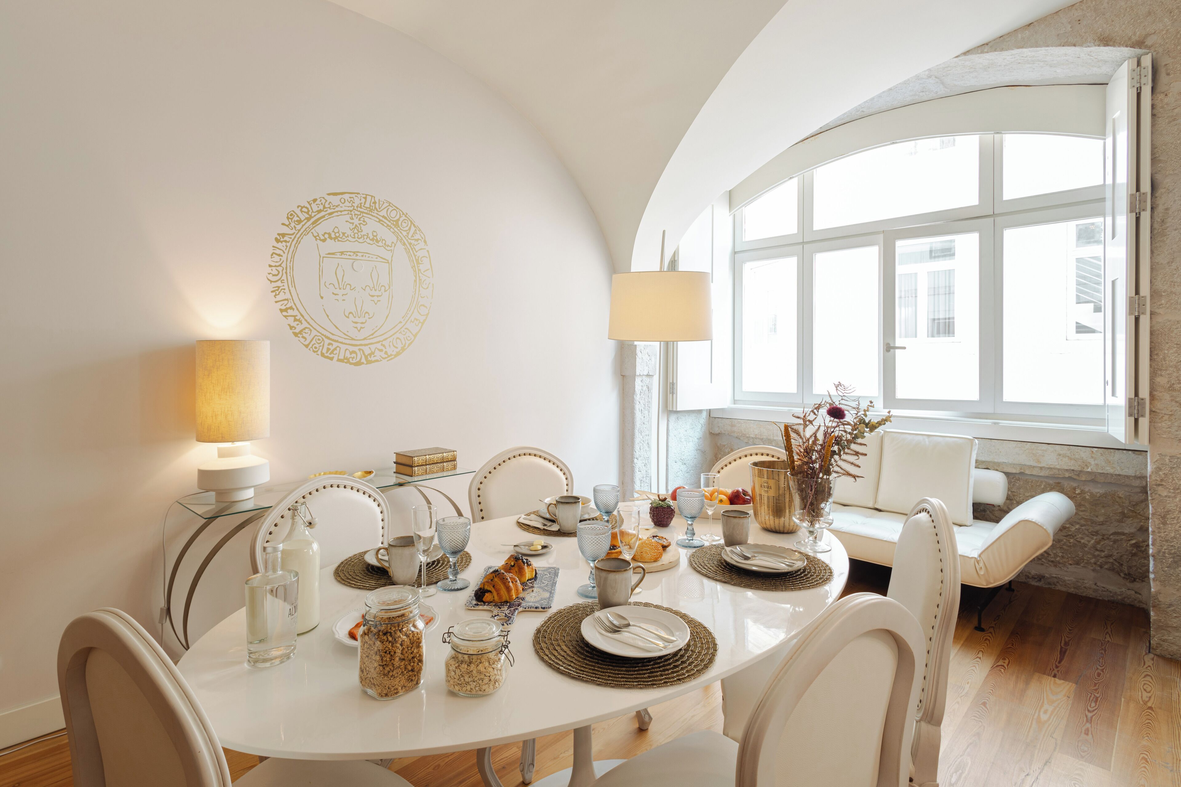 Zona pranzo con tavolo bianco apparecchiato per quattro persone, circondata da sedie bianche, una lampada e un emblema decorativo al muro.