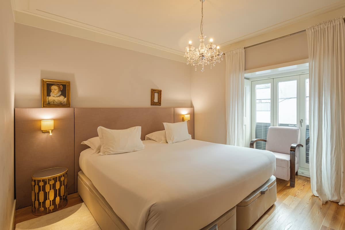 A spacious bedroom with a large bed, a chandelier, a nightstand, a chair, and a window with curtains.