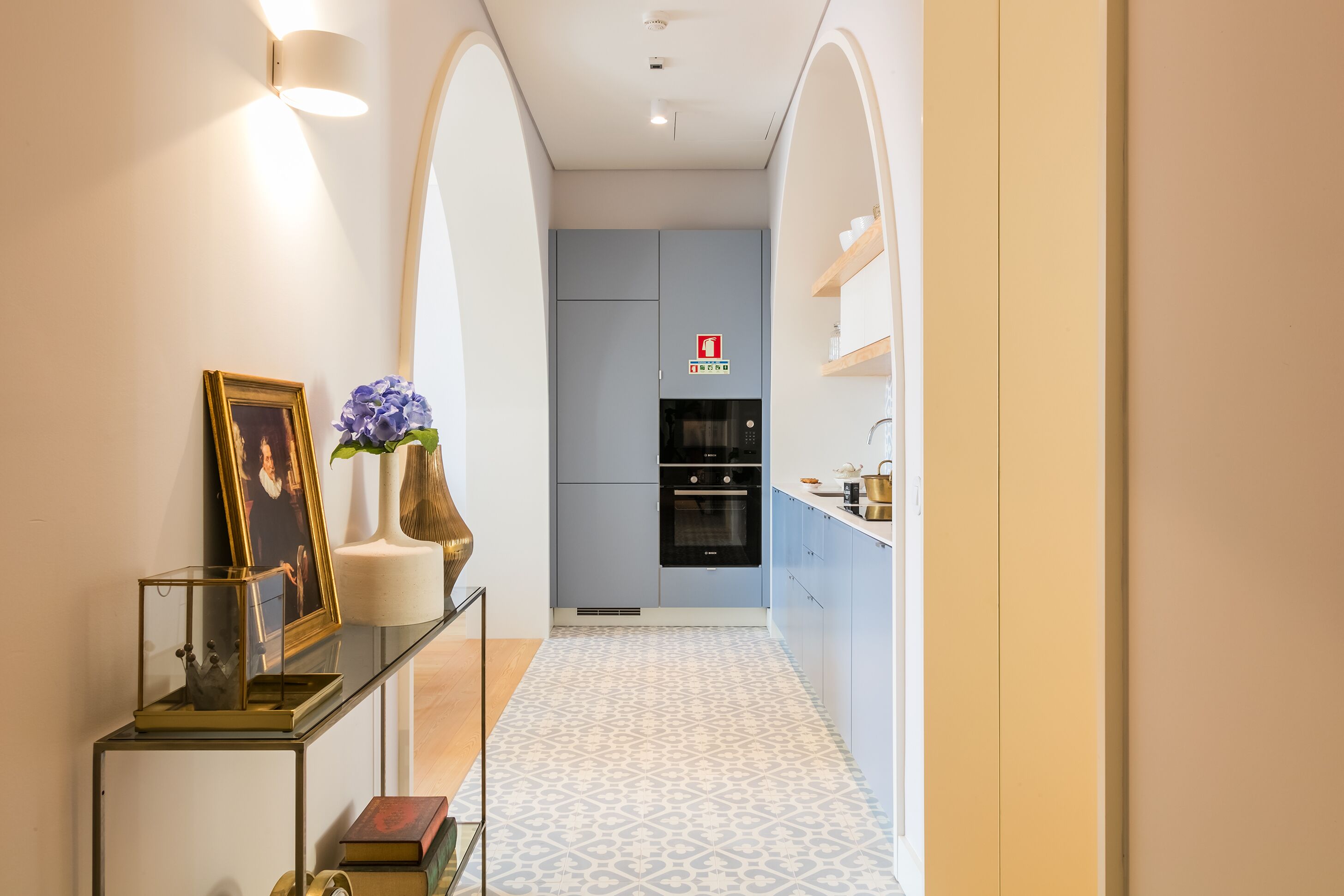 A narrow hallway leading to a kitchen with a patterned floor, a framed picture, a vase with flowers, and a fire safety sign.