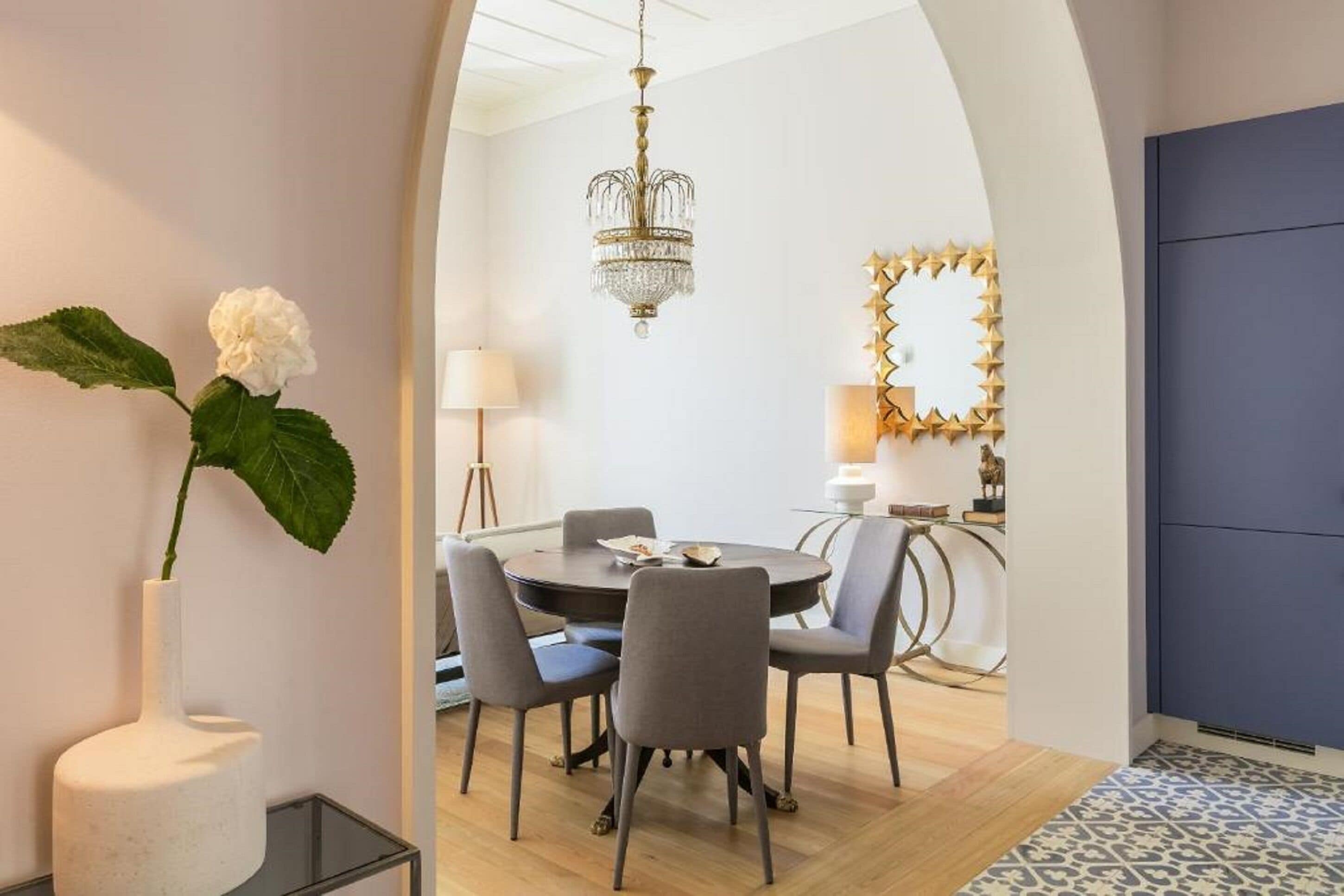 A modern dining area with a round table, grey chairs, a chandelier, and a decorative mirror.