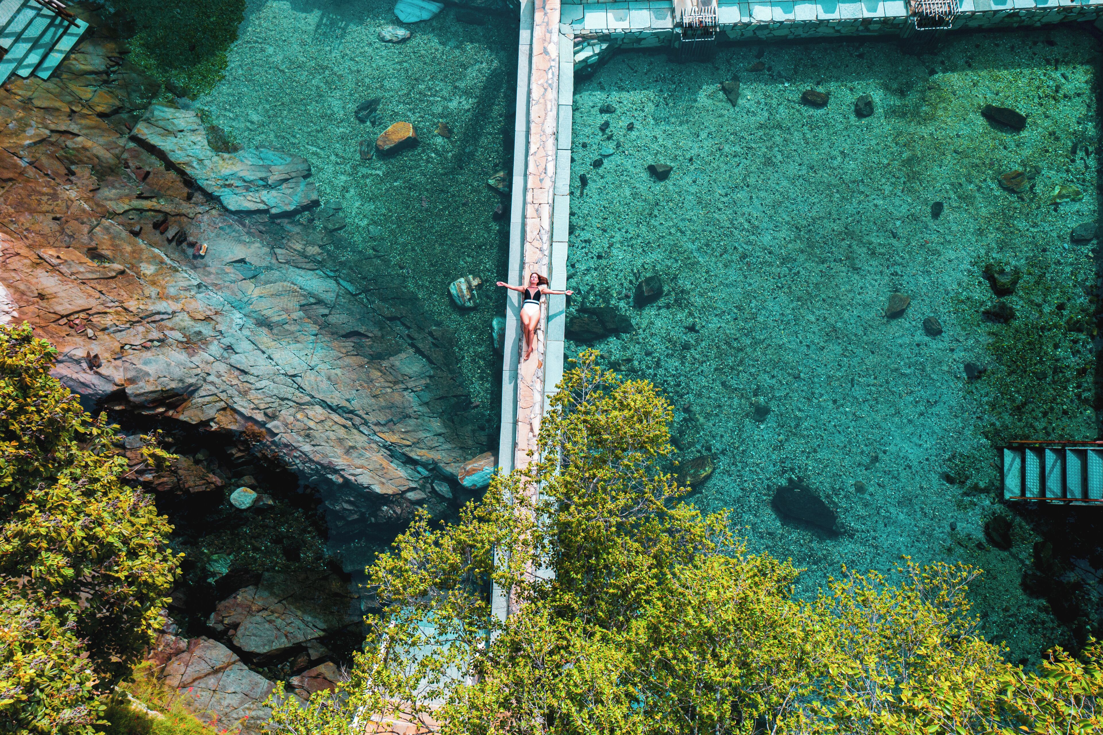 A person on a bridge over a clear water body with rocky terrain and trees.