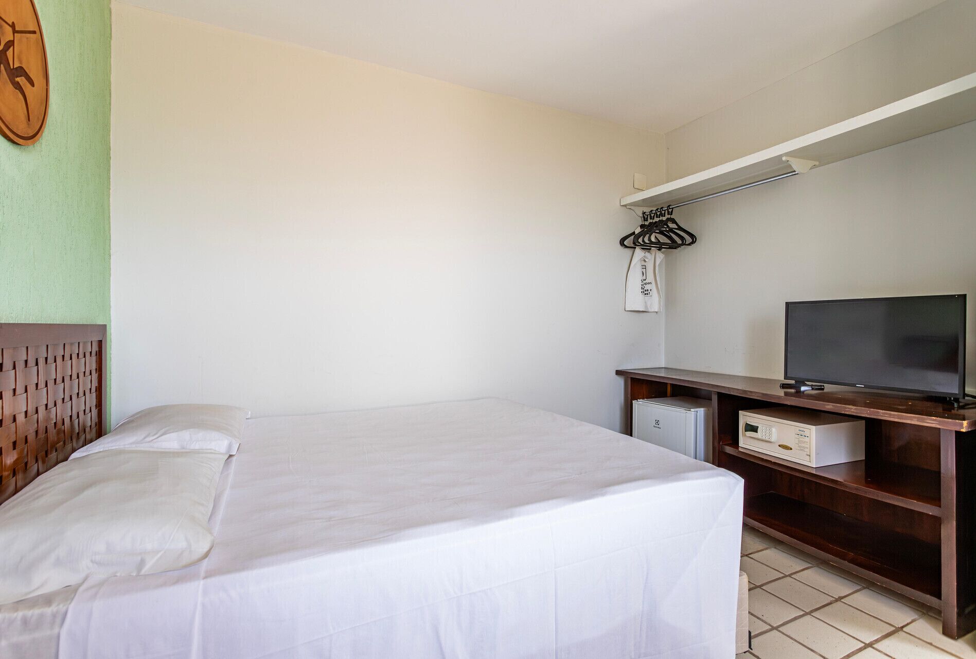 A hotel room with a bed, a television, a desk with a computer, and a wall-mounted light fixture.