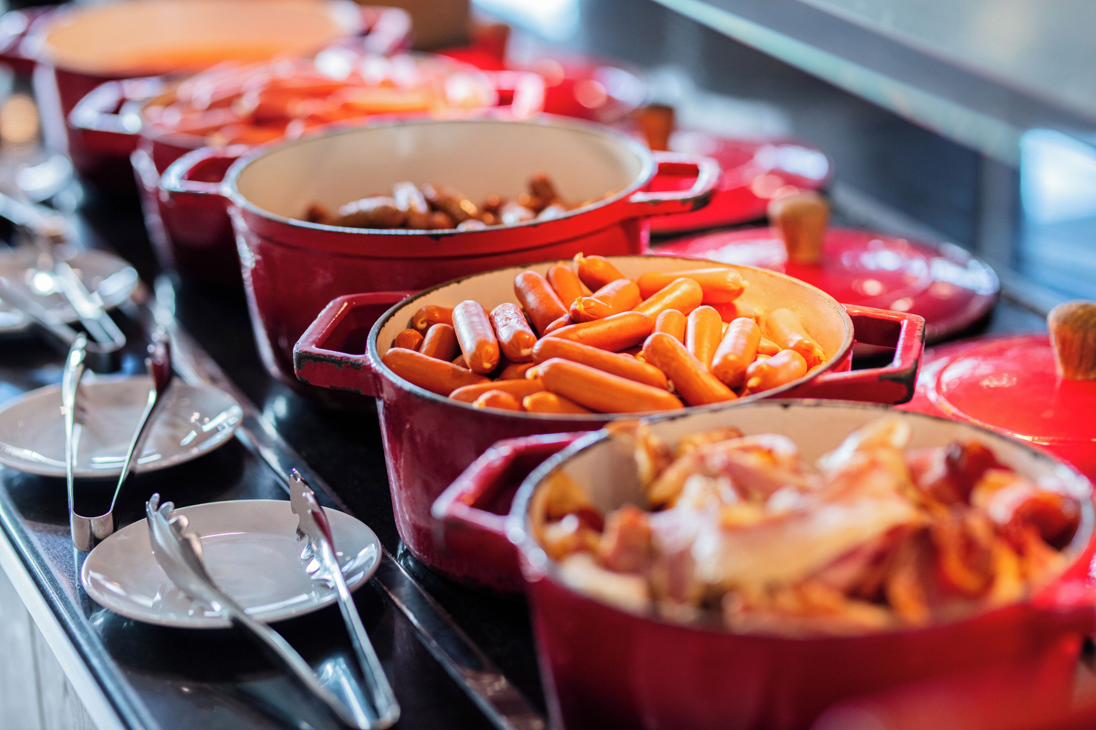 A buffet with various dishes in red pots, including carrots and sausages.