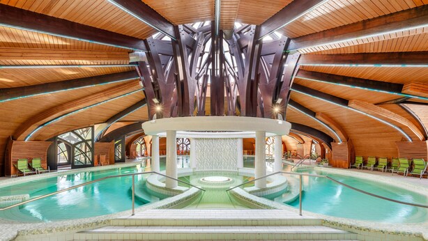 Una amplia piscina interior con una fuente central, rodeada por una estructura de madera con techos abovedados y sillas verdes a lo largo del borde.