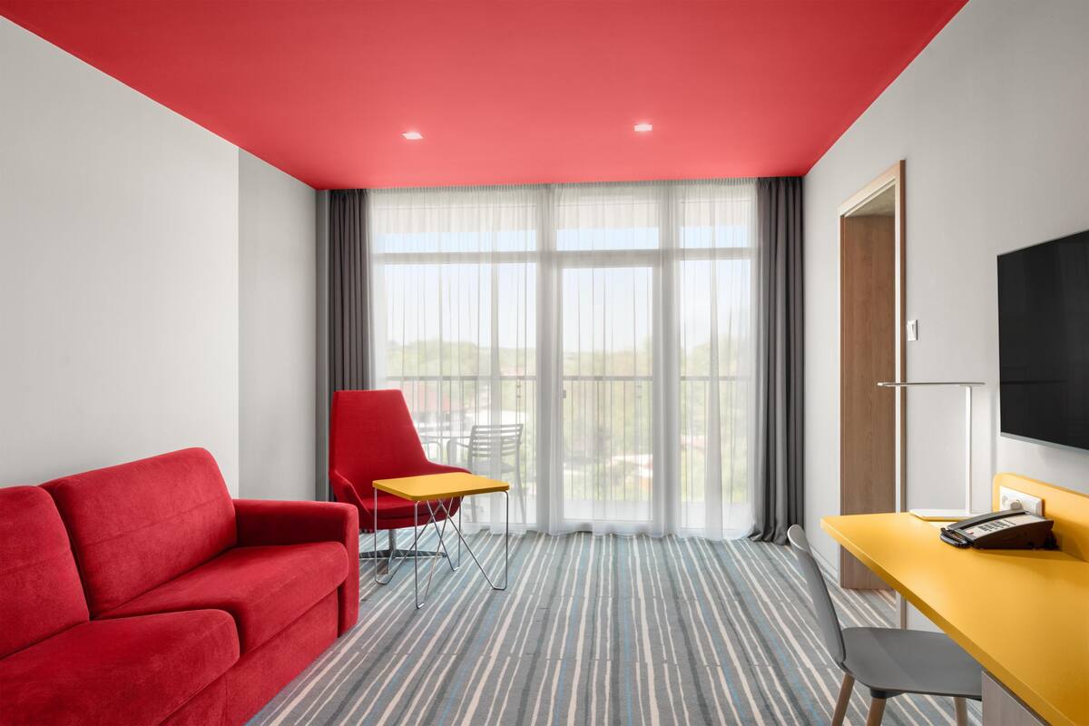 Habitación de hotel con techo rojo, sofá rojo, escritorio amarillo, silla roja y una ventana grande con cortinas.
