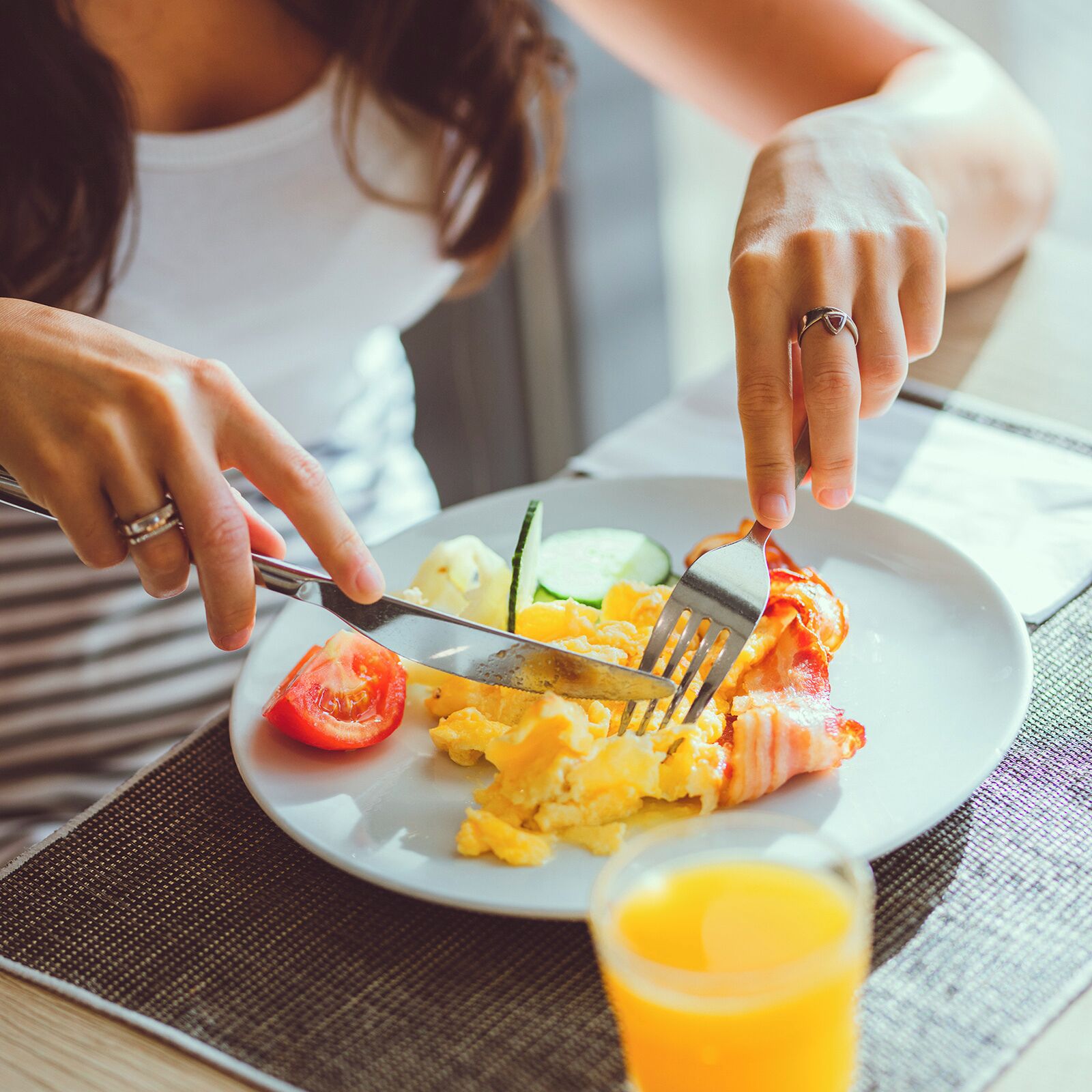 A person eating scrambled eggs with bacon, a tomato, and a cucumber on a plate.