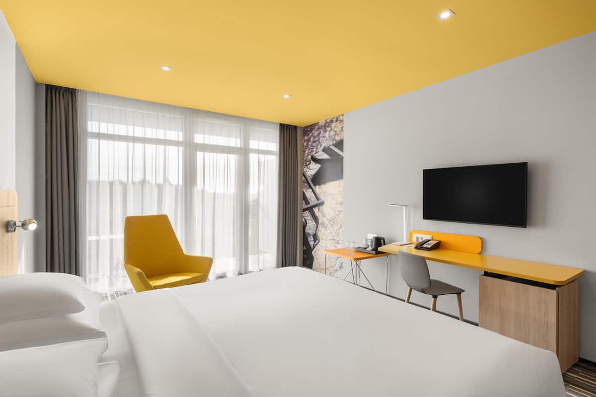 Una habitación de hotel con cama, escritorio, silla, televisor y ventana con cortinas.