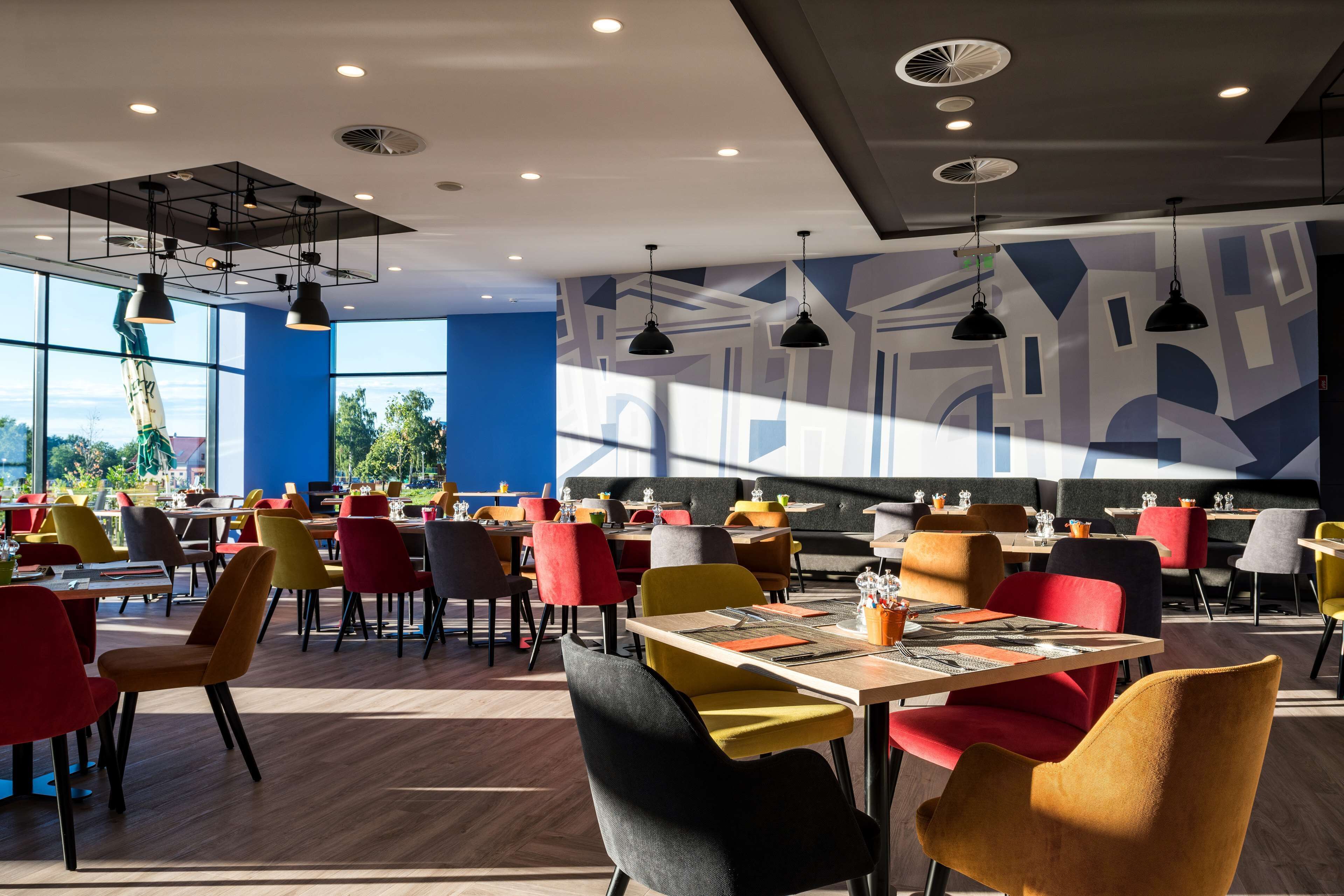 Ein modernes Restaurant mit bunten Stühlen und Tischen, großen Fenstern und geometrischer Wandkunst.