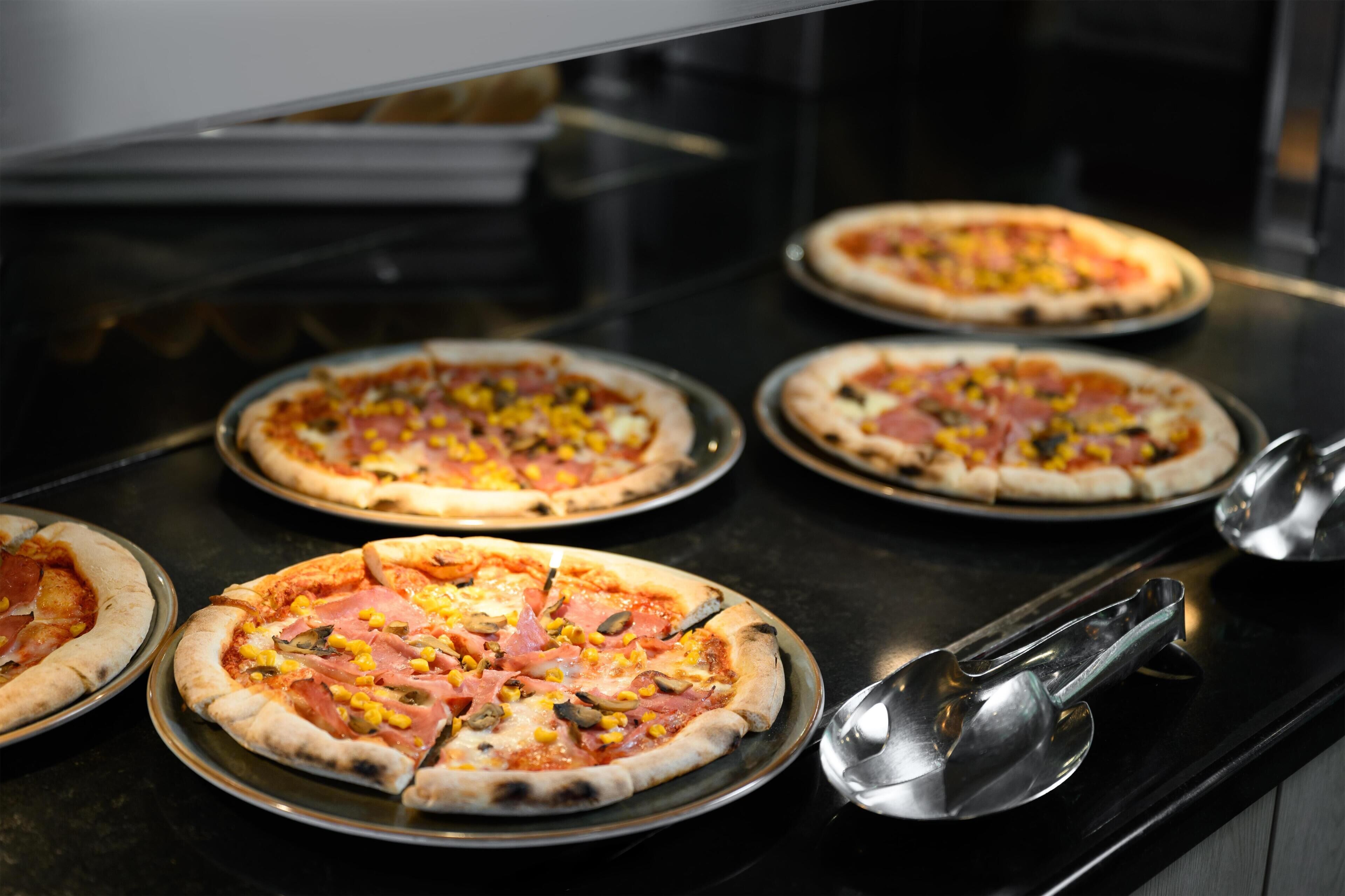 Eine Küchenarbeitsplatte mit mehreren Pizzen auf Metalltellern, einem Metallschöpfkelle und einem Pizzaschneider.