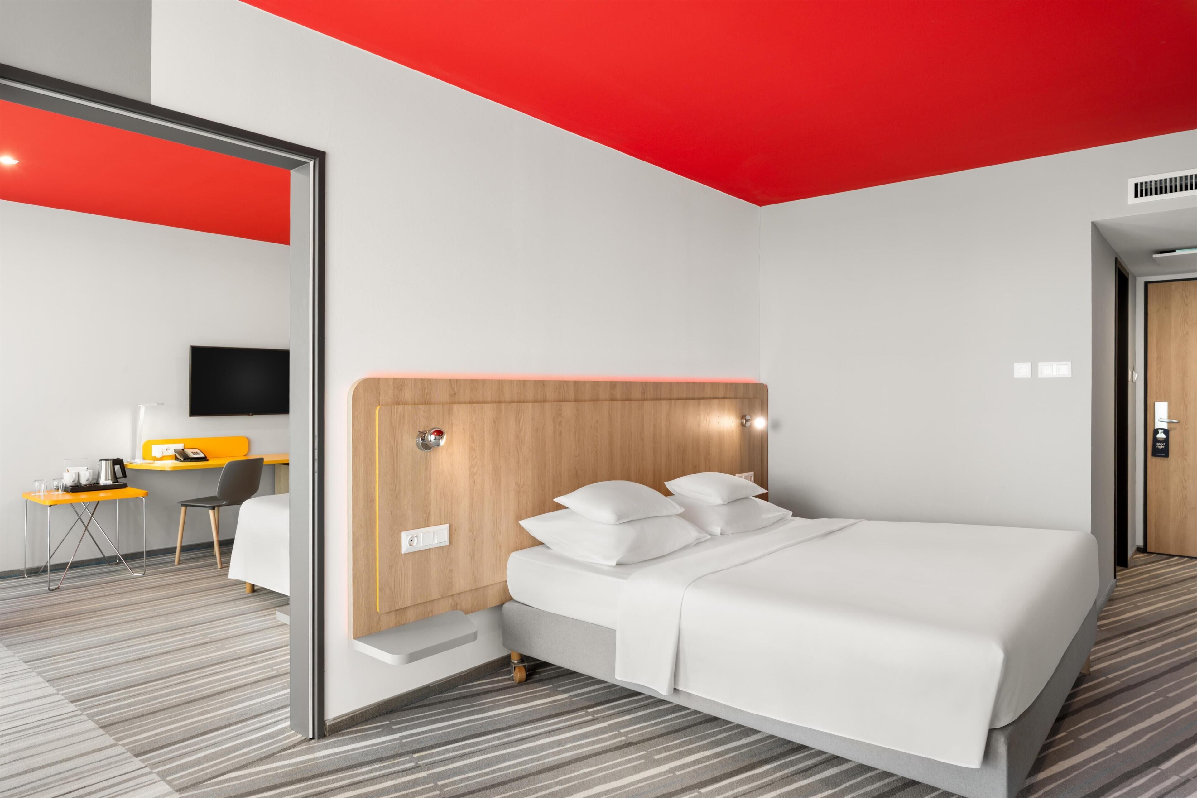 A modern hotel room with a bed, a desk with a chair, a television, and a red accent wall.