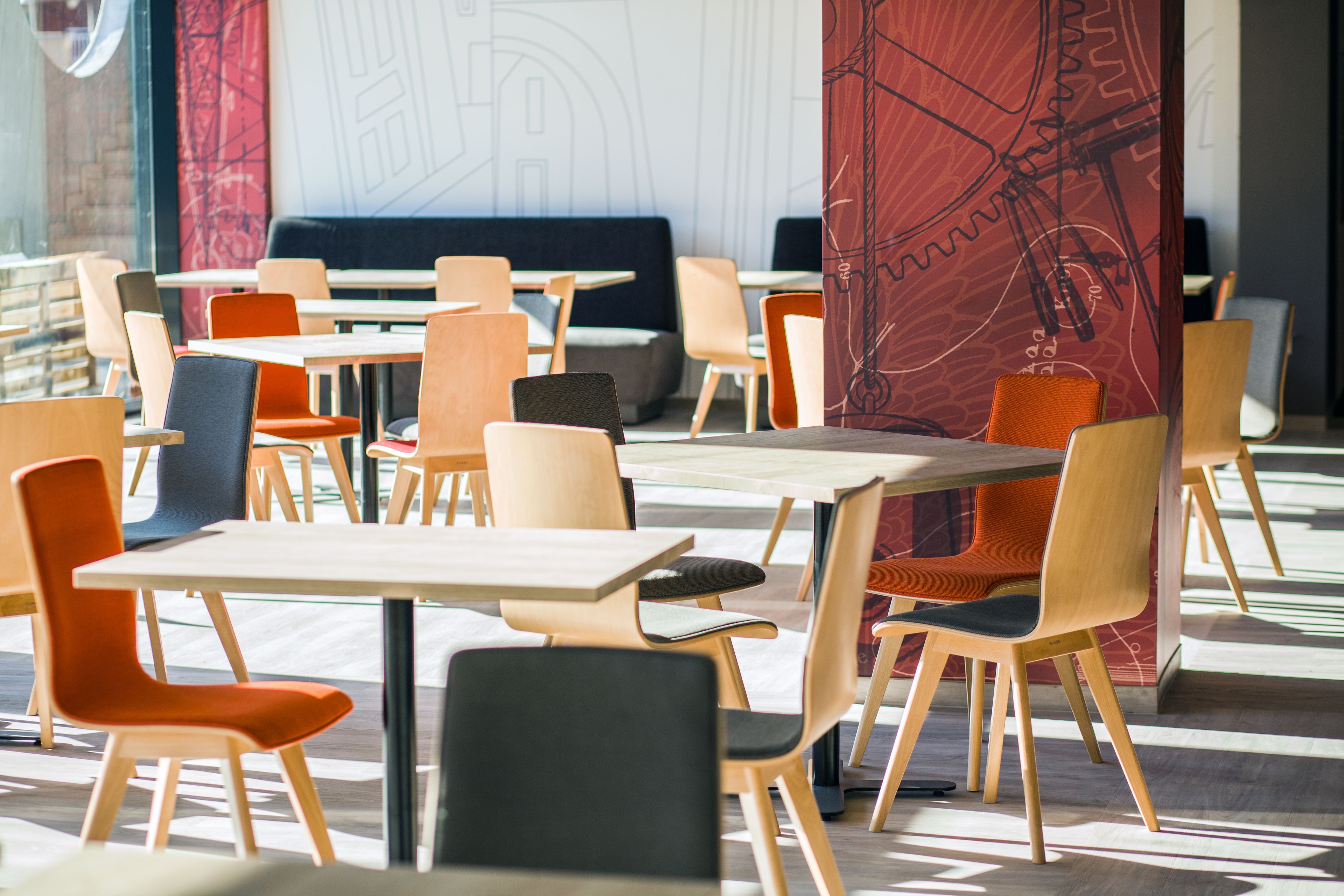 A modern dining area with multiple tables and chairs, a large window, and a red abstract wall design.