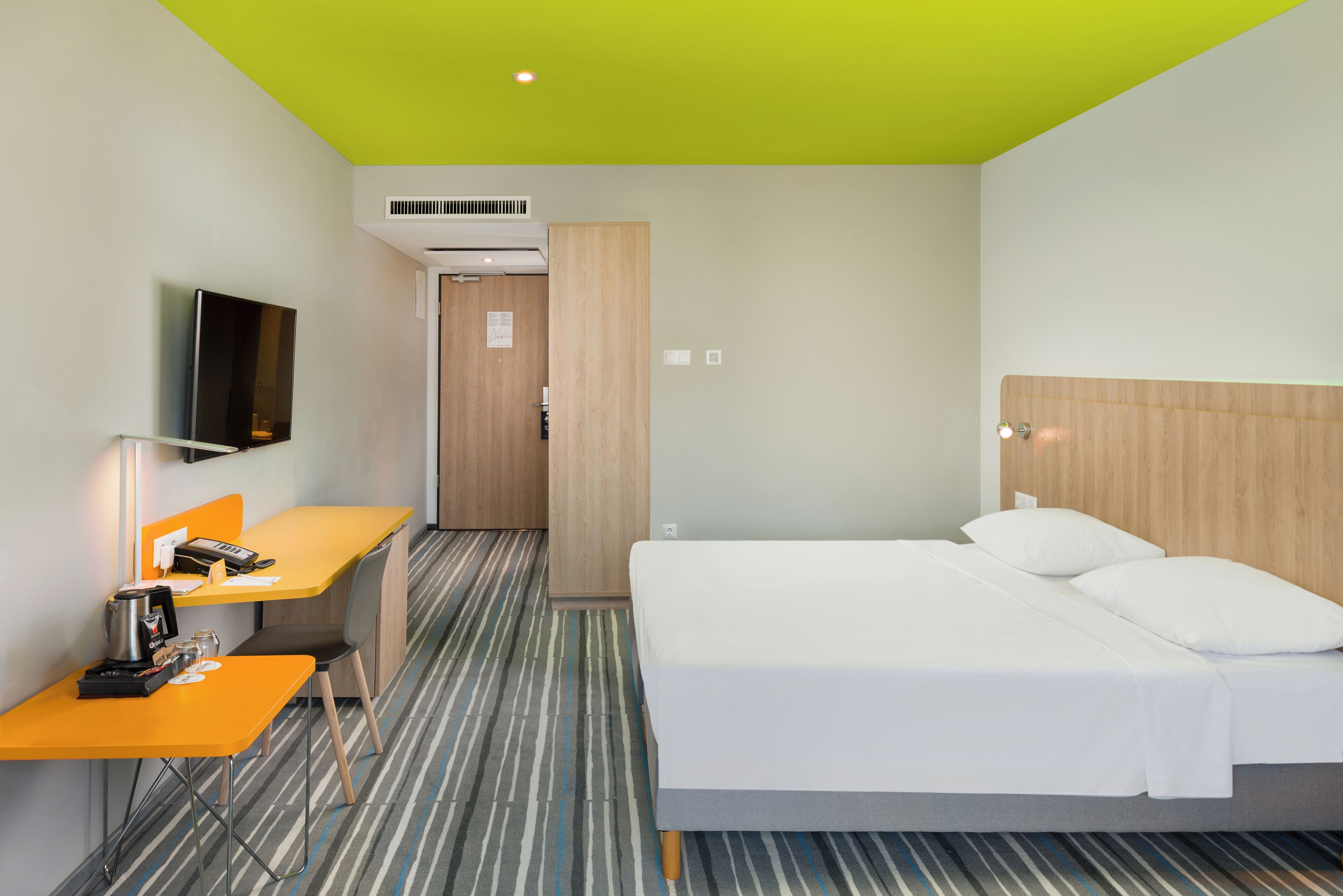 A hotel room with a bed, a desk with a coffee maker, a television, and a door leading to another room.
