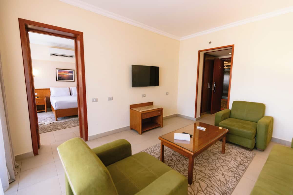 A hotel room with a bed, a TV mounted on the wall, a green sofa, a wooden coffee table, and a patterned rug.