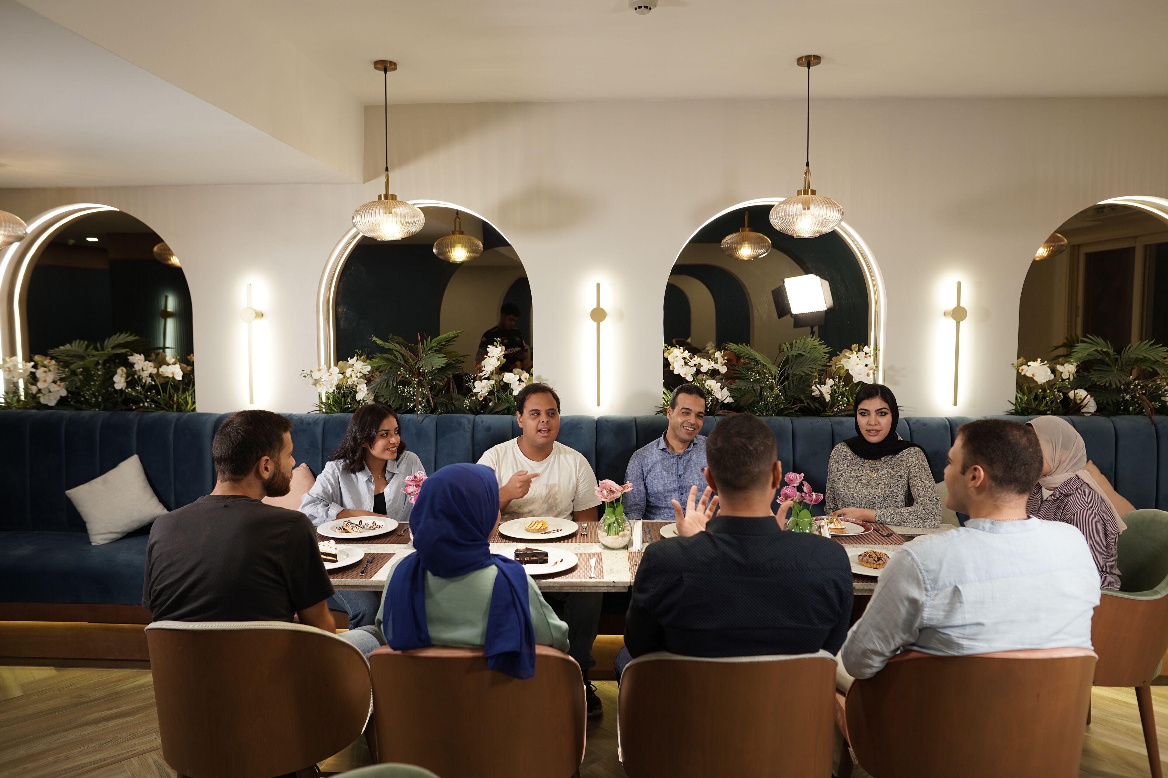 A group of people dining at a restaurant with a modern interior, featuring a blue upholstered seating area and pendant lighting.