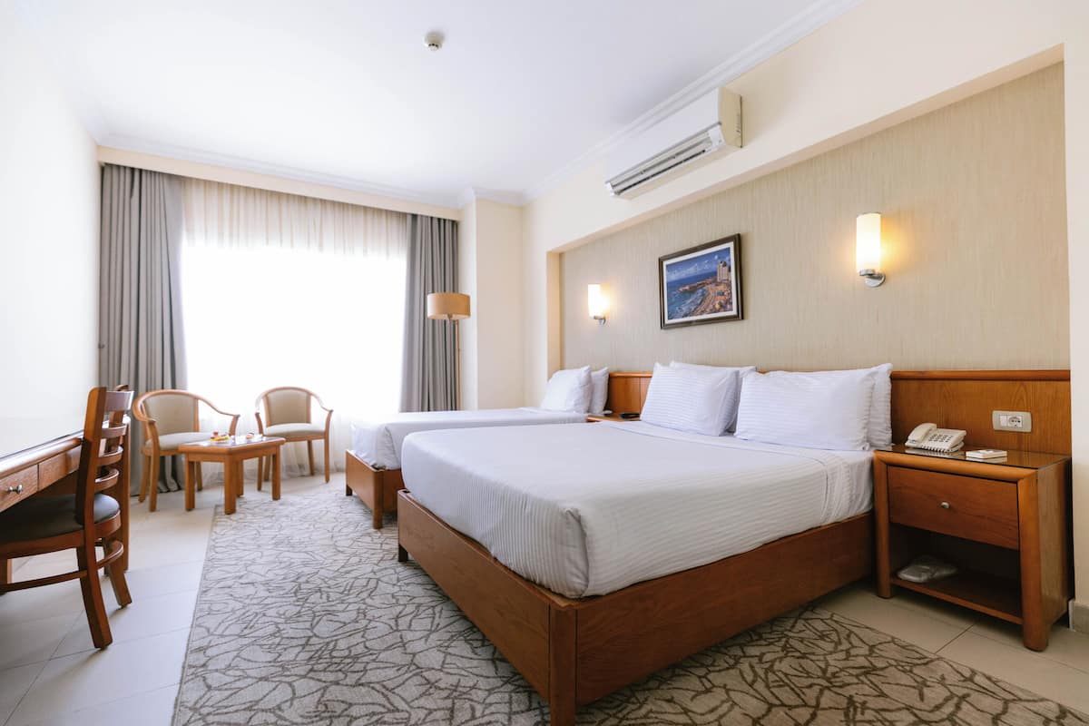 A hotel room with a large bed, two bedside tables, a desk with a chair, a small table with chairs, and a large window with curtains.