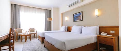 A hotel room with a large bed, two bedside tables, a desk with a chair, a small table with chairs, and a large window with curtains.