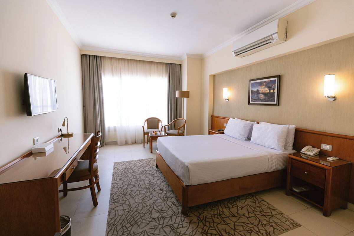 A hotel room with a bed, desk, chairs, a TV, and a painting on the wall.