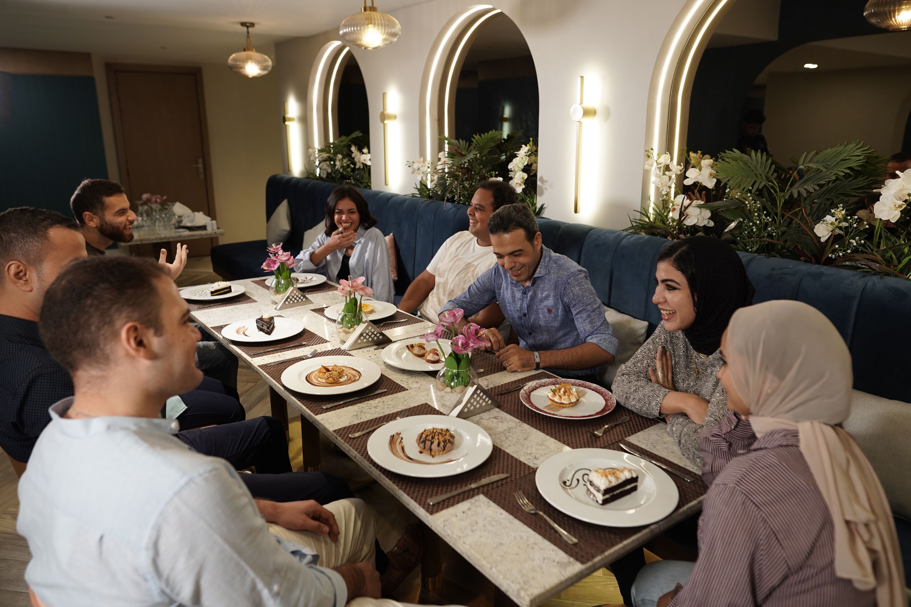 A group of people seated at a long table with plates of food, in a modern interior with arched lighting and floral decorations.