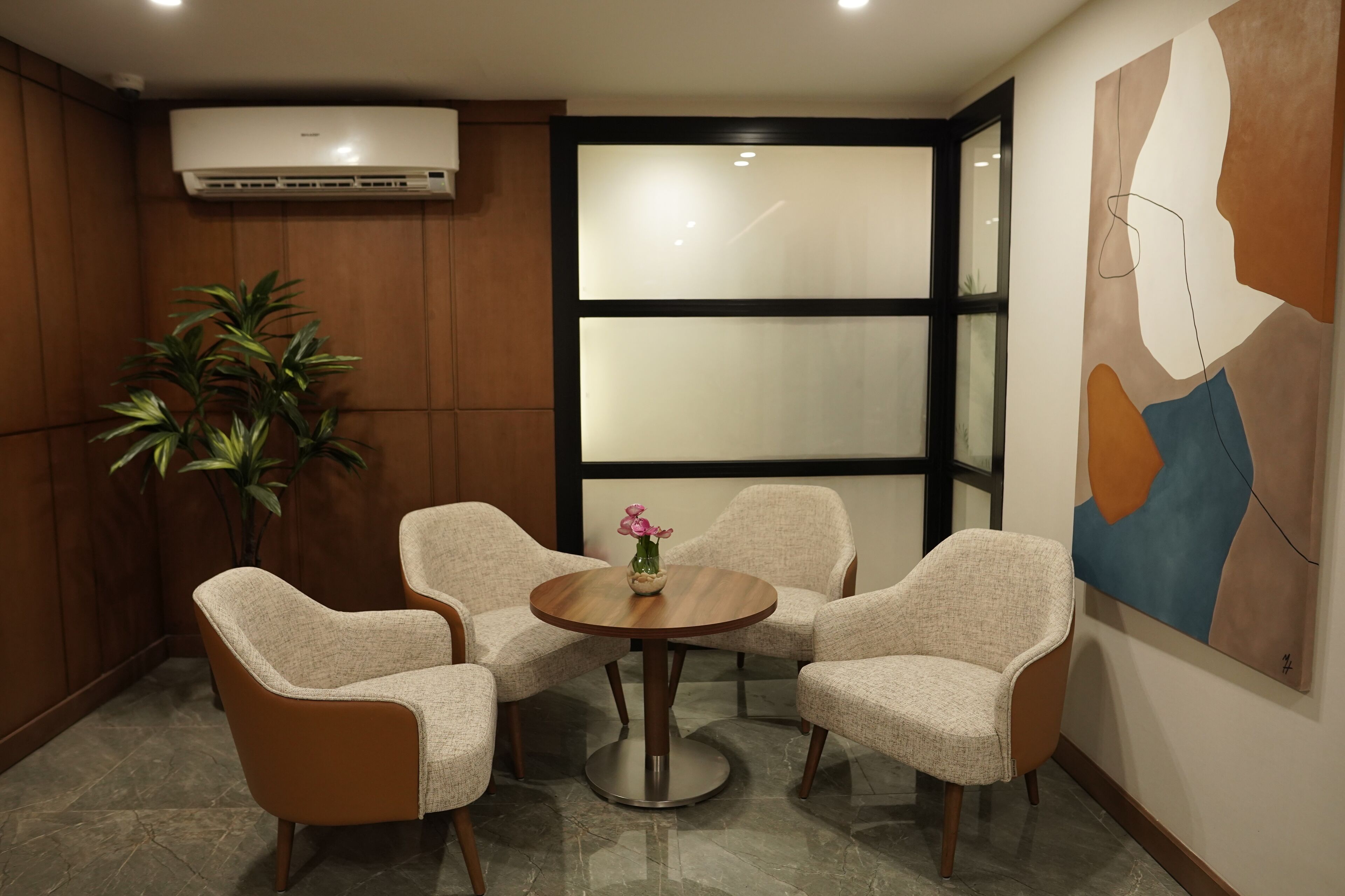 Un salón de conferencias con cuatro sillas color arena alrededor de una mesa redonda, una planta en maceta y un cuadro abstracto grande en la pared.
