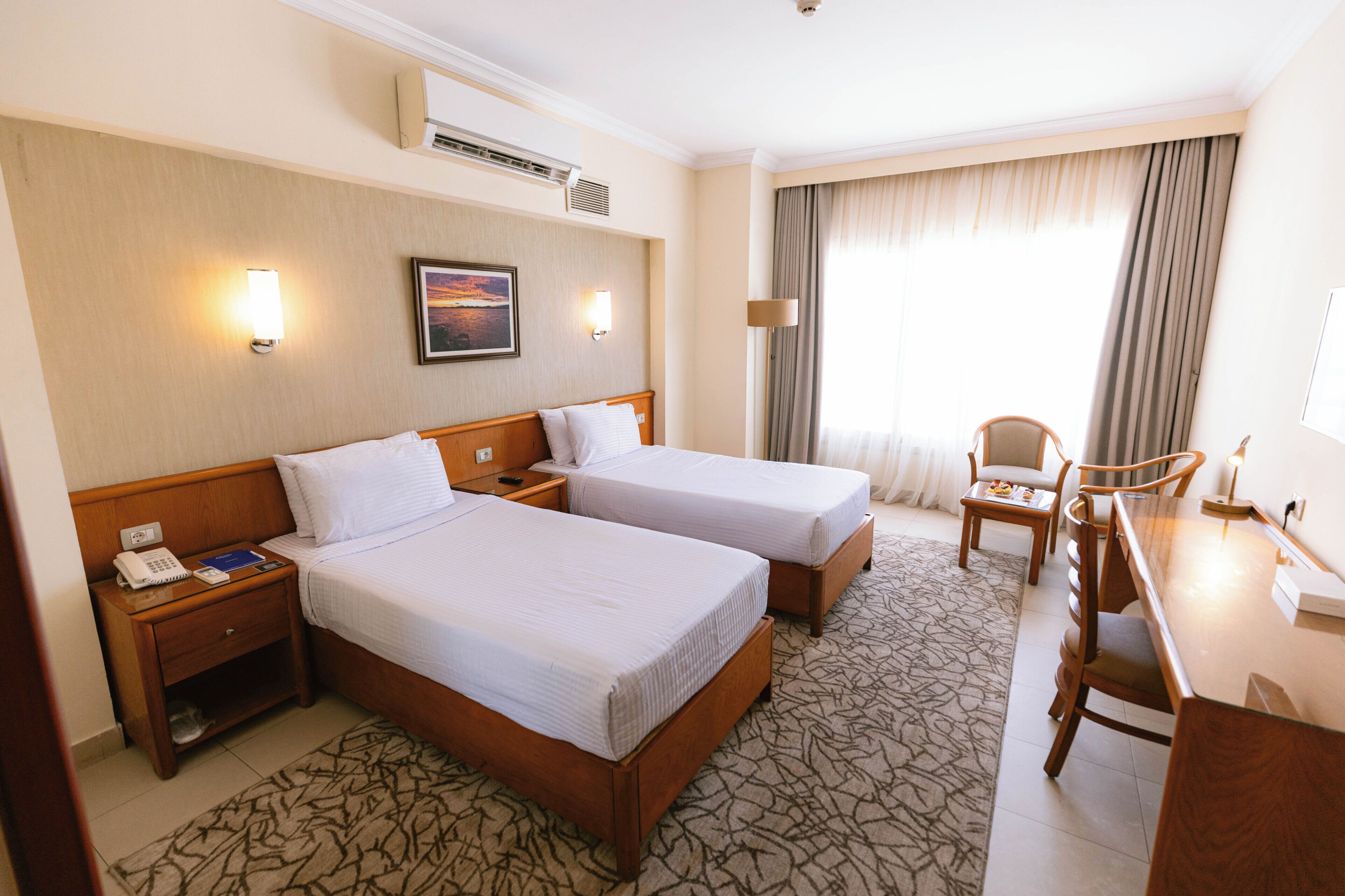 A hotel room with two beds, a desk, a chair, a nightstand with a phone, and a wall-mounted air conditioner.