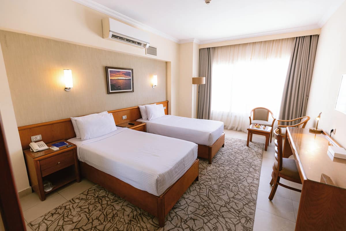 A hotel room with two beds, a desk, a chair, a nightstand with a phone, and a wall-mounted air conditioner.