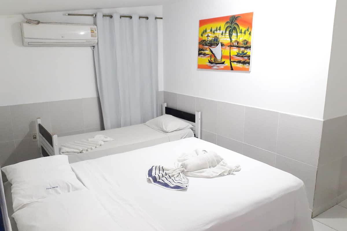 A hotel room with two single beds, white bedding, a painting of a sunset scene, and an air conditioning unit on the wall.