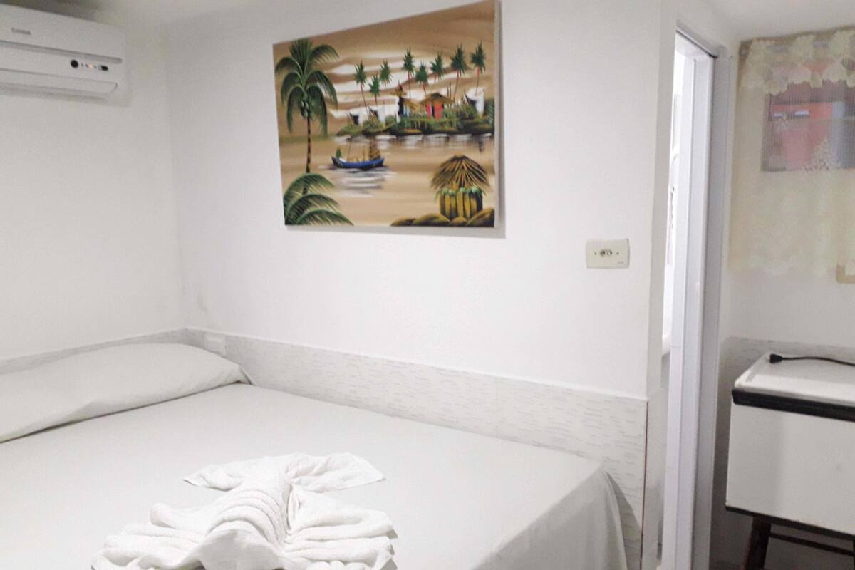 A hotel room with a bed, a painting of a beach scene, a white desk, and a white chair.