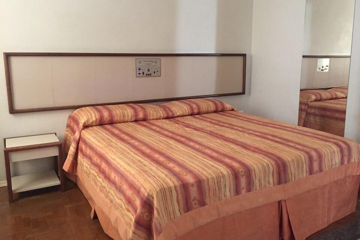 A single-bed room with a striped bedspread, a nightstand, a mirror, and a power outlet on the wall.