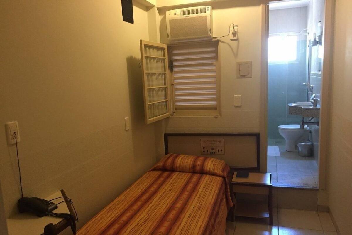 A small, compact room with a single bed, a bedside table, a window with blinds, a radiator, and a view of a bathroom through an open door.