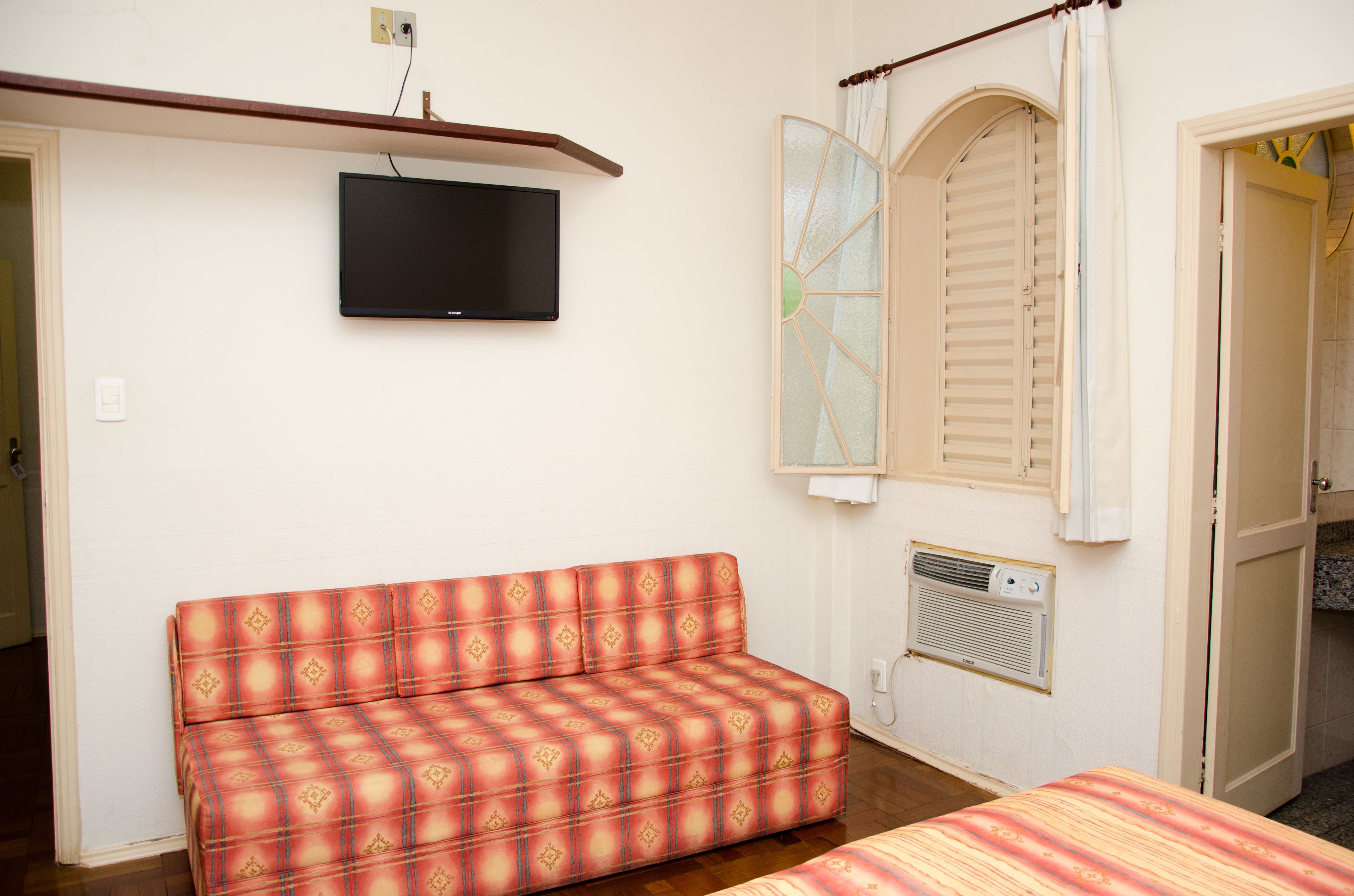 Ein Zimmer mit einer Couch, einem an der Wand montierten Fernseher, einem Fenster mit Rollläden und einer Tür zu einem anderen Raum.